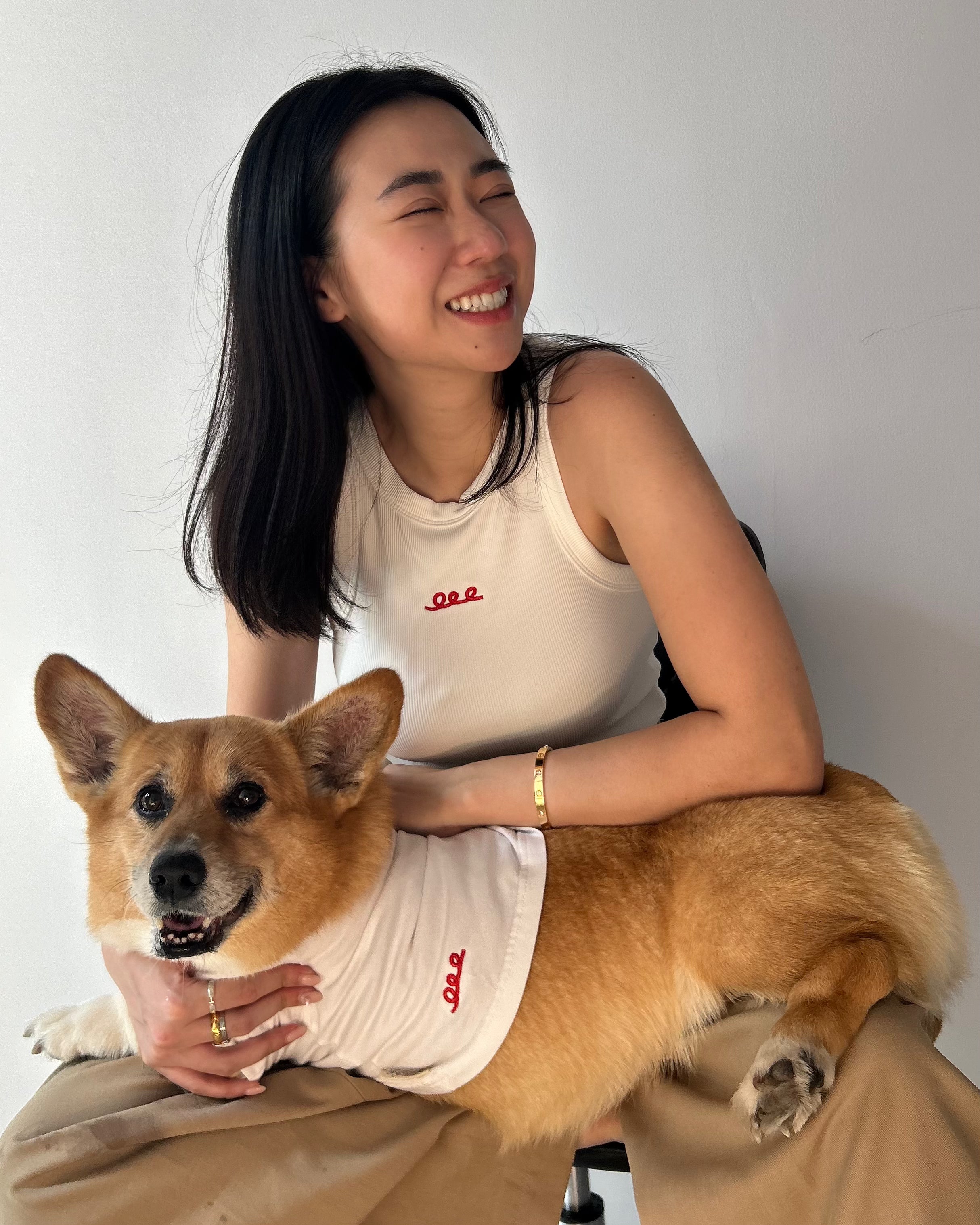 Woman sitting with a corgi dog wearing matching white shirts against a plain background