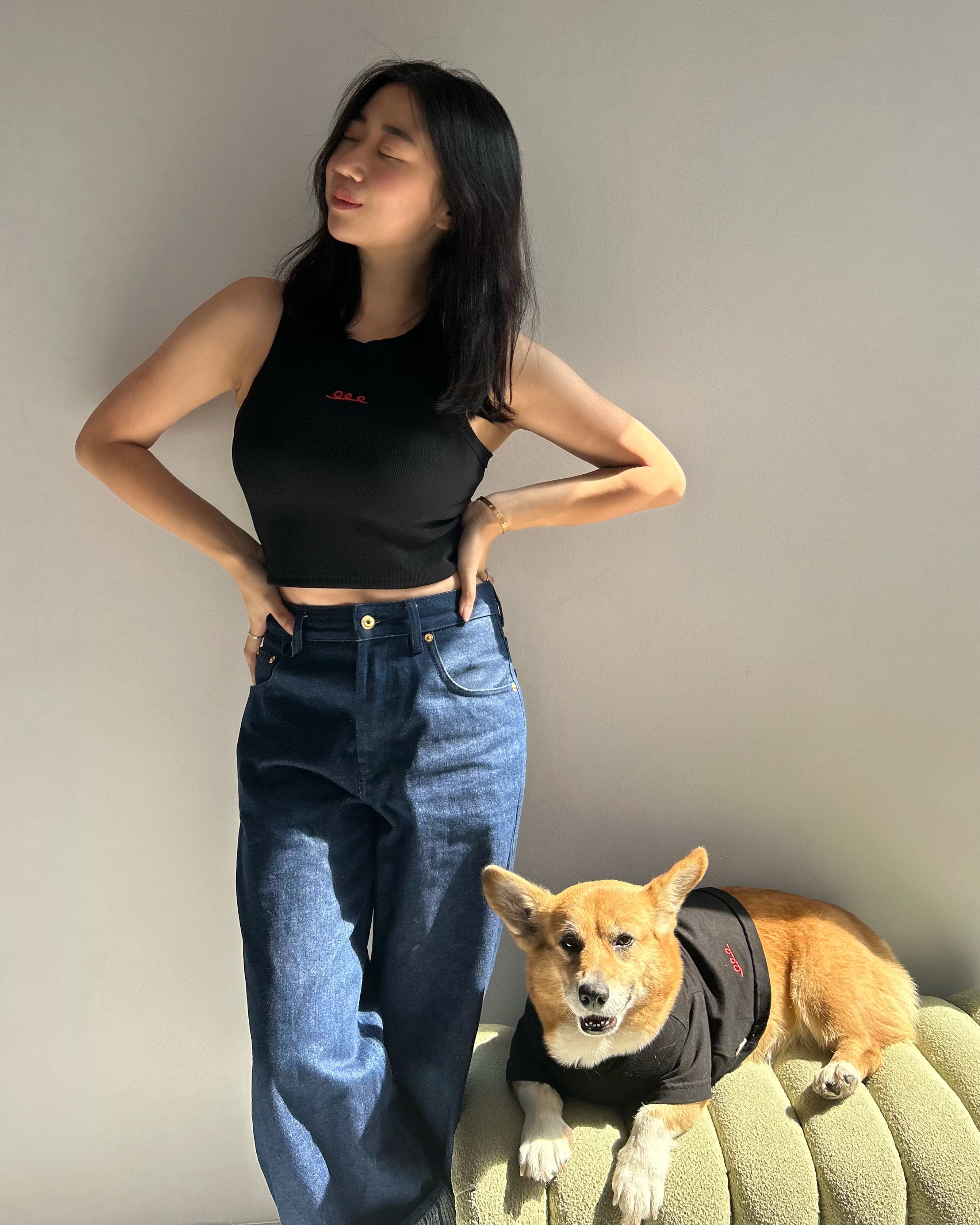Woman in black top and blue jeans standing next to a dog on a couch.