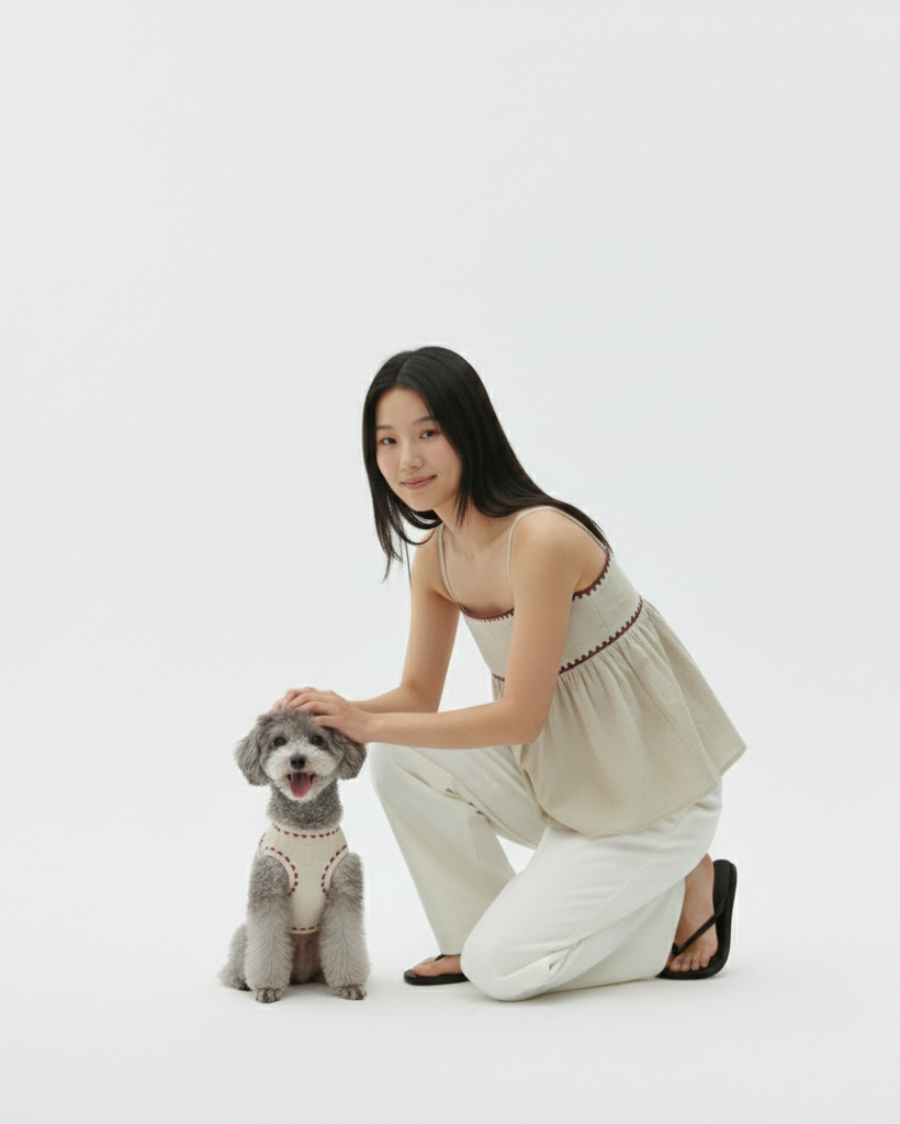 Woman in a beige top and white pants kneeling next to a small dog on a white background
