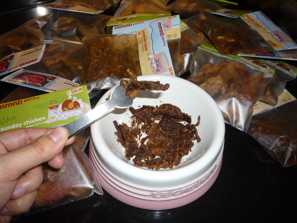 Dried chicken pieces in a container with a spoon, surrounded by packages on a dark surface.