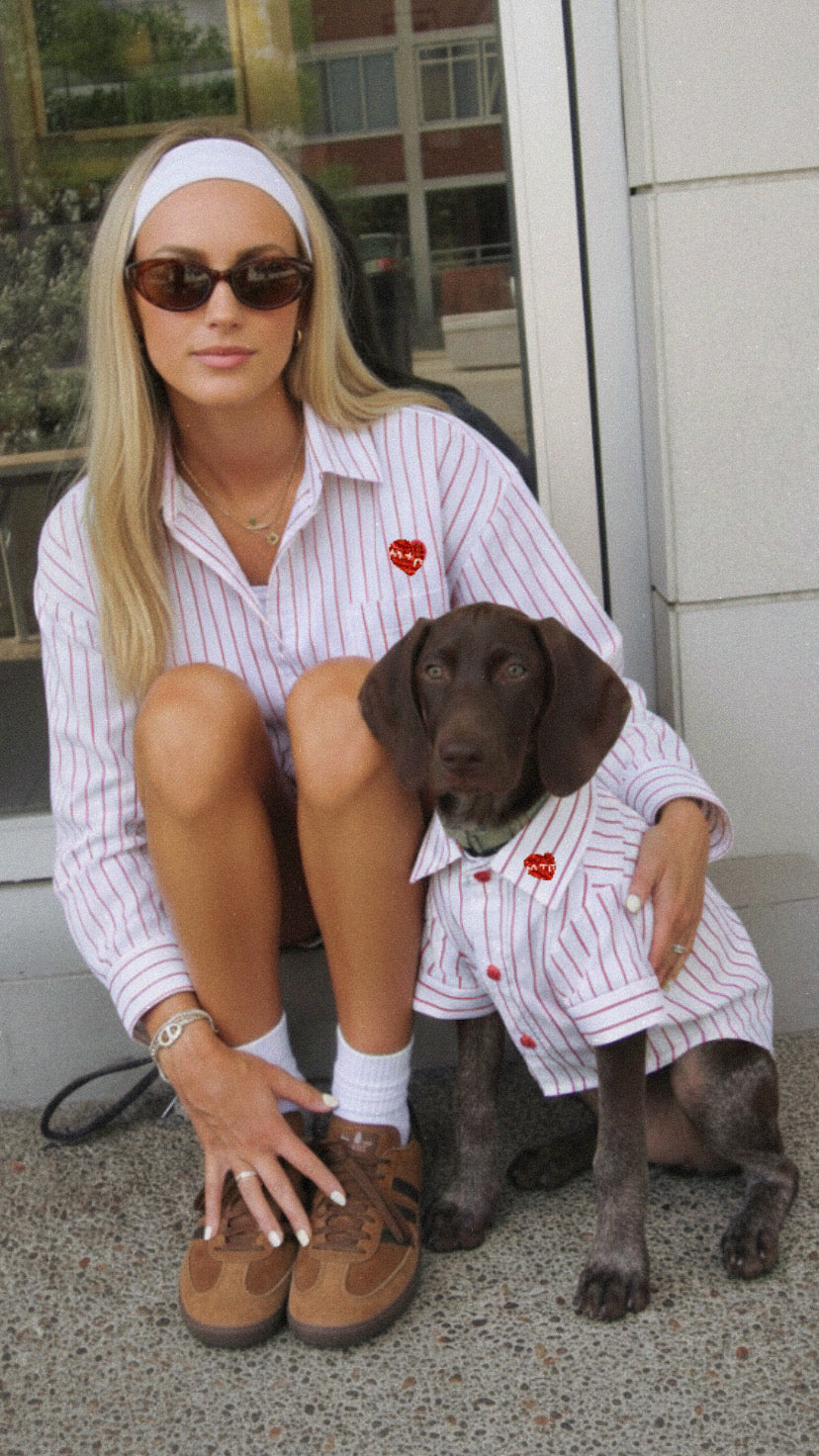 Woman in a striped shirt and sunglasses with a dog wearing a matching outfit, sitting on a sidewalk.